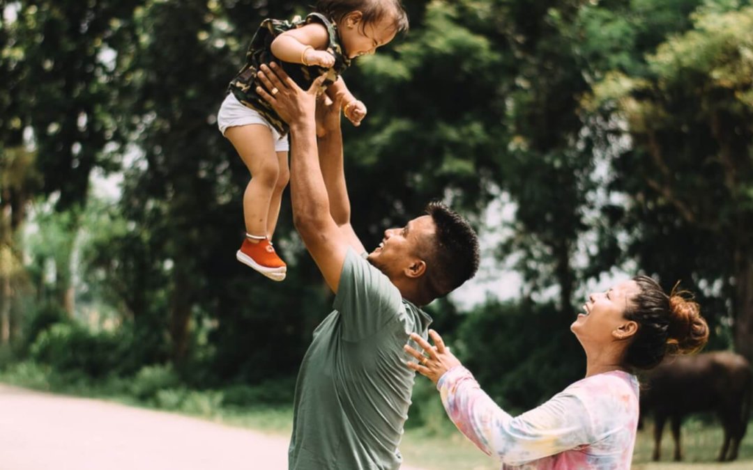 A dad plays with a child and holds him high in the air, while a mom watches and smiles.