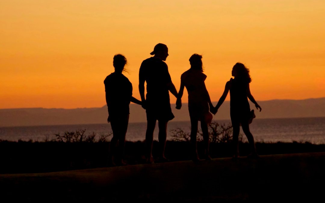 A family of two parents and two children stands silhouetted against an orange sunset